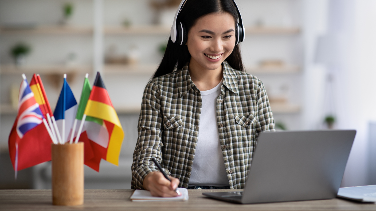 Happy young asian woman learning foreign language online from home using wireless headset and making notes